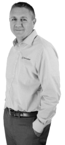 A black and white image of Adam Jackson from the waist up smiling with his hand in his pocket while wearing a Power/mation long sleeve button up shirt
