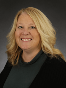 Sue Clarin smiling for a photo from the chest up while wearing a dark gray shirt with a black cardigan